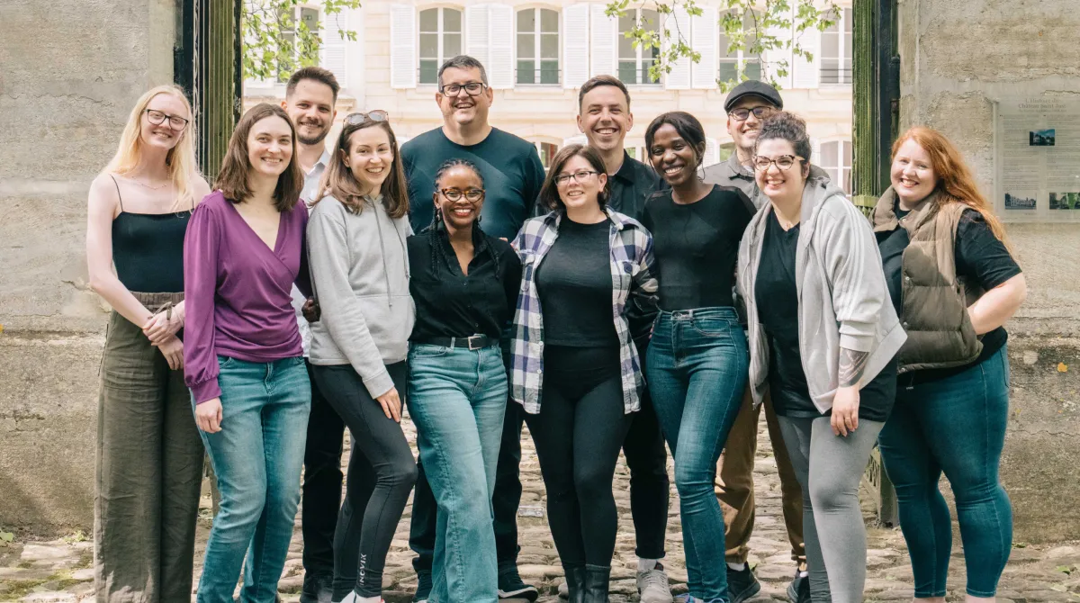 A diverse group of twelve smiling people standing closely outside a building under tree branches.