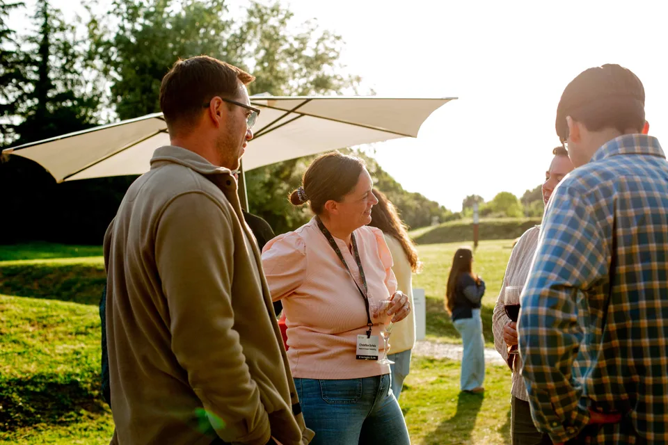 A sunlit group conversation under a white umbrella, a teammate in pink laughing